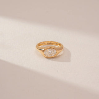 Close-up view of a bezel-set diamond ring in 14KT Yellow Gold, featuring an oval centre diamond and a sculpted band against a beige backdrop.