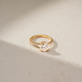 Side view of a round solitaire engagement ring in 14 KT Yellow Gold, featuring a round lab-grown diamond in a four-prong setting with a pavé-set diamond band, displayed against a beige backdrop.