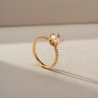 Angled view of a round solitaire engagement ring in 14 KT Yellow Gold, featuring a round lab-grown diamond in a four-prong setting with a pavé-set diamond band, displayed against a beige backdrop.
