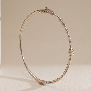 A close-up side angle of the Basic Solitaire Diamond Bracelet in 14KT hallmarked white gold. The sleek, minimalist bangle features a textured beaded band with a bezel-set round solitaire diamond at the center. The bracelet is displayed standing upright with its spring lock clasp visible, placed against a soft beige background that enhances the metallic shine