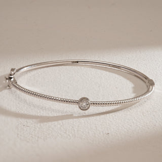 Overhead view of the Basic Solitaire Diamond Bracelet crafted in white gold with hallmarked 14KT purity. The bracelet’s fine beaded detailing runs across the band, drawing attention to the bezel-set solitaire diamond centerpiece. Its refined spring lock clasp design can be partially seen, while the neutral beige surface provides a subtle backdrop highlighting the bracelet’s symmetry and elegance.