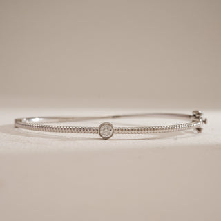 Front-facing close shot of the Basic Solitaire Diamond Bracelet in 14KT white gold. The bracelet’s slim, textured band is accentuated by a single bezel-set round solitaire diamond in the center. Photographed on a beige surface, the image captures the delicate craftsmanship, polished metal finish, and timeless elegance of this minimalist diamond bangle.