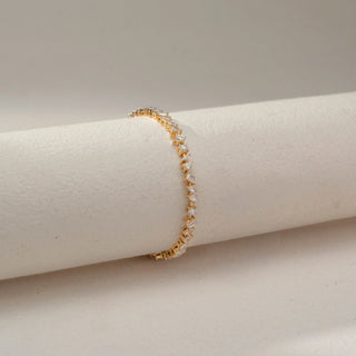 Side view of a mixed-shape diamond tennis bracelet in 14 KT Dual Gold, featuring alternating emerald-cut, pear-shaped, and round lab-grown diamonds in elevated settings, displayed on a beige surface.
