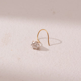 Angled view of a yellow gold nose pin, featuring a solitaire round diamond in a minimal prong setting. Captured against a soft beige backdrop, the design reflects delicate sparkle and understated elegance.
