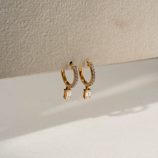 Side view of diamond hoop earrings in yellow gold, featuring pavé-set diamonds and dangling emerald-cut diamond charms. Displayed against a neutral backdrop, the profile highlights elegant craftsmanship and movement.