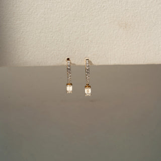 Front view of diamond drop earrings in yellow gold, featuring pavé-set diamond bars accented with dangling emerald-cut diamonds. Displayed against a neutral backdrop, the design offers sleek elegance.