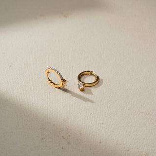 Angled view of yellow gold diamond hoop earrings with pavé-set stones and dangling emerald-cut diamond charms, displayed on a soft neutral surface with natural light and shadows.