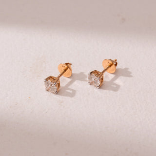 Angled view of round diamond stud earrings in yellow gold, featuring solitaire diamonds in four-prong settings. Captured against a soft beige backdrop, the design reflects timeless sparkle and elegance.