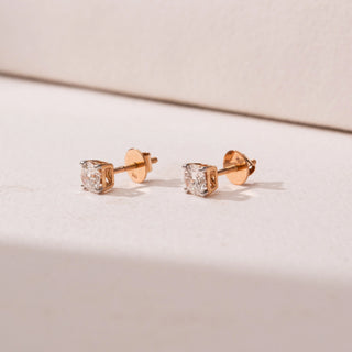 Side view of round diamond stud earrings in yellow gold, featuring solitaire diamonds in four-prong settings. Captured against a soft beige backdrop, the design reflects timeless sparkle and elegance.