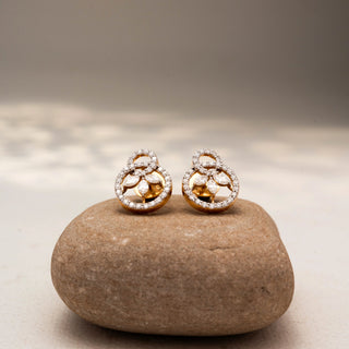 Front view of floral diamond stud earrings in 14 KT Yellow Gold, featuring pavé-set circular motifs with marquise diamond petals, displayed on a textured stone surface.
