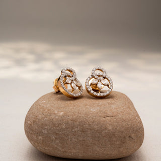 Angled view of floral diamond stud earrings in 14 KT Yellow Gold, featuring pavé-set circular motifs with marquise diamond petals, displayed on a textured stone surface.
