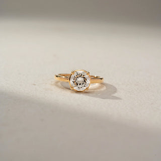 Front view of a round solitaire engagement ring in 14 KT Yellow Gold, featuring a round lab-grown diamond in a classic four-prong setting on a sleek polished band, displayed against a beige backdrop.