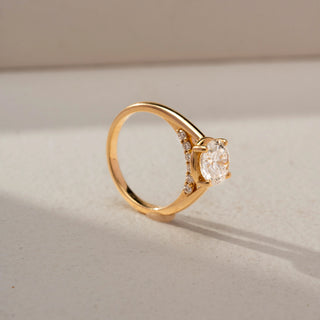 Angled view of a round solitaire engagement ring in 14 KT Yellow Gold, featuring a round lab-grown diamond in a classic four-prong setting on a sleek polished band, displayed against a beige backdrop.