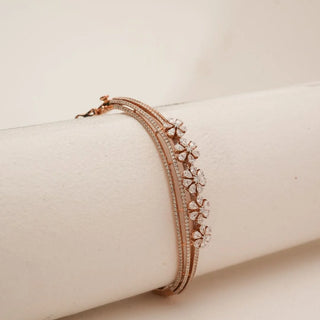 Angled view of rose gold floral diamond bangle showcasing layered blossom motifs with pear-shaped petal diamonds and pavé-set bands, resting on a soft neutral display surface.