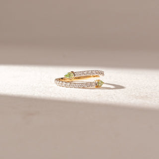 Front view of open wrap ring in yellow gold with two pear-shaped green gemstones at each end and pavé diamond bands, set on a soft neutral surface, highlighting vibrant contrast and delicate sparkle.
