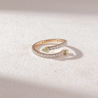Top view of open wrap ring in yellow gold with two pear-shaped green gemstones and pavé diamond bands, set on a soft neutral surface, highlighting elegant curves and fresh sparkle.
