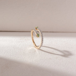 Angled view of open wrap ring in yellow gold with two pear-shaped green gemstones and a single pavé diamond band, set on a soft neutral background, highlighting sleek curves and delicate sparkle.
