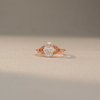 Front view of an oval solitaire engagement ring in 14 KT Rose Gold, featuring an oval-cut lab-grown diamond accented by intricate leaf-inspired details along the band, displayed against a beige backdrop.