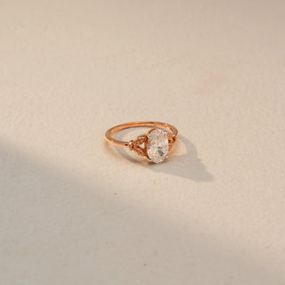 Side view of an oval solitaire engagement ring in 14 KT Rose Gold, featuring an oval-cut lab-grown diamond accented by intricate leaf-inspired details along the band, displayed against a beige backdrop.