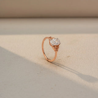 Angled view of an oval solitaire engagement ring in 14 KT Rose Gold, featuring an oval-cut lab-grown diamond accented by intricate leaf-inspired details along the band, displayed against a beige backdrop.