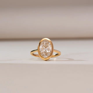 Front view of Modern Bezel Solitaire Diamond Ring in yellow gold, featuring an oval lab-grown diamond secured in a sleek bezel setting, displayed on a soft beige backdrop.
