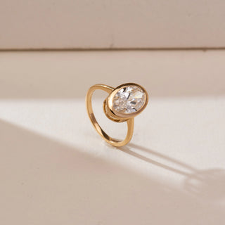 Angled view of Modern Bezel Solitaire Diamond Ring in yellow gold, showcasing an oval lab-grown diamond secured in a smooth bezel setting, displayed on a soft beige backdrop.