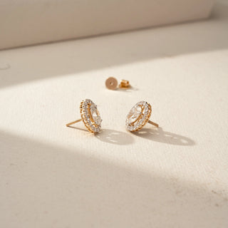 Angled view of oval-cut diamond halo stud earrings in yellow gold, showcasing the sparkling halo setting, elevated profile, and secure threaded posts on a neutral surface.