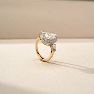Angled view of a pear halo engagement ring in 14 KT Yellow Gold, featuring a pear-shaped lab-grown diamond encircled by a double halo and accented by pear-shaped side diamonds in elevated settings, displayed against a beige backdrop.