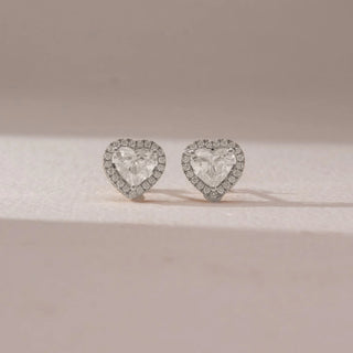 Front view of diamond stud earrings in 14KT yellow gold, featuring heart-shaped center diamonds surrounded by pavé halo settings, displayed on a beige surface highlighting romantic elegance and sparkle.