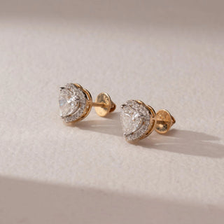 Side view of diamond stud earrings in 14KT yellow gold, featuring heart-shaped center diamonds with pavé halo settings and screw backs, displayed on a beige surface highlighting depth and brilliance.
