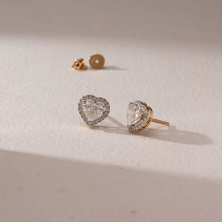 Angled view of diamond stud earrings in 14KT yellow gold, featuring heart-shaped center diamonds with pavé halo settings and screw backs, placed on a beige surface highlighting detail and sparkle.