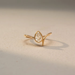 Front view of a pear solitaire ring in 14 KT Yellow Gold, featuring a pear-shaped lab-grown diamond nestled within a sleek sculpted bypass setting, displayed against a beige backdrop.