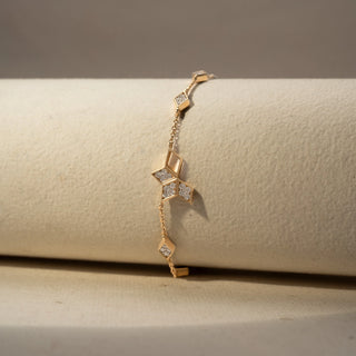 Close up  view of a geometric diamond bracelet in 14 KT Yellow Gold, featuring open kite-shaped motifs accented with clustered diamonds and delicate chain links, displayed against a soft beige backdrop.
