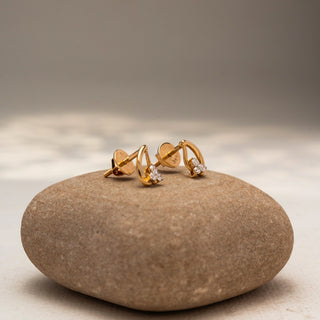Side view of diamond stud earrings in 14 KT Yellow Gold, featuring a trio of round diamonds nestled within sleek curved gold motifs, displayed on a textured stone surface.
