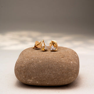 Angled view of diamond stud earrings in 14 KT Yellow Gold, featuring a trio of round diamonds nestled within sleek curved gold motifs, displayed on a textured stone surface.