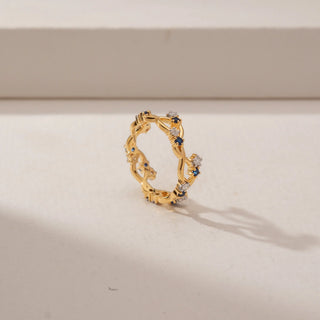 Angled view of a blue sapphire and diamond ring in 14KT Yellow Gold, featuring an intertwined vine band against a beige backdrop.