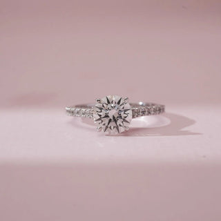 Front view of Elegant Pavé Shank Diamond Ring in white gold, showcasing a round lab-grown centre diamond secured in prongs with pavé-set diamonds along a slender band on a soft pink surface.