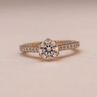 Front view of Reia’s 14 KT Yellow Gold Pure Elegance Ring. The centred round solitaire and its shimmering pavé diamond band are showcased against a soft pink surface.