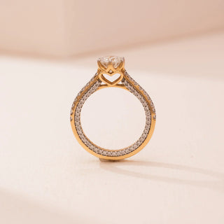 Side view of Reia’s 14 KT Yellow Gold Pure Elegance Ring. The round solitaire is held in a heart-shaped four-prong setting on a shimmering pavé diamond band against a soft pink surface.