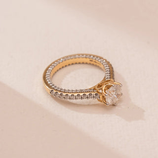 Top view of Reia’s 14 KT Yellow Gold Pure Elegance Diamond Solitaire Ring. A round solitaire sits in a four-prong setting on a pavé diamond band against a soft pink surface.