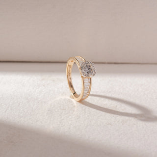 Angled view of round diamond solitaire ring in yellow gold with baguette diamond accents on the band, set on a soft neutral background, highlighting sleek detail and radiant sparkle.
