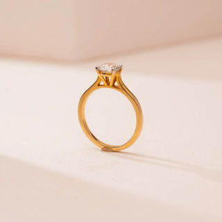 Side view of Reia’s 14 KT Yellow Gold Classic Crown Set Solitaire Ring. The round diamond is held high in a crown setting on a smooth gold band, shown on a soft pink surface.