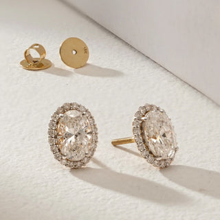 Oval-cut lab-grown diamond halo stud earrings in 18KT Yellow Gold, featuring EF-VVS diamonds with a radiant halo design, displayed on a soft neutral surface with Bombay screw backs.