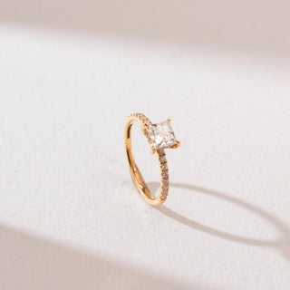 Angled view of a 14KT yellow gold princess-cut solitaire diamond ring with a square center stone in four prongs and a slim pavé diamond band, set against a soft neutral background.