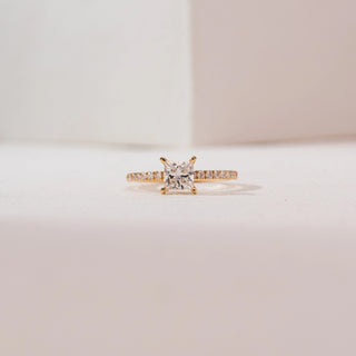 Front view of a 14KT yellow gold princess-cut solitaire diamond ring with a square center stone in four prongs and a slim pavé diamond band, set against a soft neutral background.