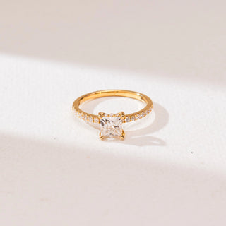 Top view of a 14KT yellow gold princess-cut solitaire diamond ring with a square center stone in four prongs and a slim pavé-set diamond band on a soft neutral surface.
