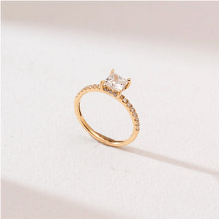 Side angle view of a 14KT yellow gold princess-cut solitaire diamond ring with a square centre stone in four prongs and a slim pavé diamond band, set on a soft neutral background.