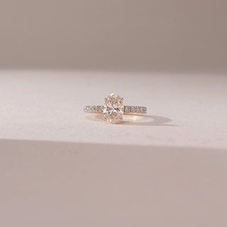 Front view of oval diamond solitaire ring in yellow gold with a pavé diamond band, set on a soft neutral surface, highlighting elongated shape and radiant, light-catching sparkle.
