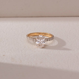 Top view of oval diamond solitaire ring in yellow gold with a pavé diamond band, set on a soft neutral surface, highlighting elegant proportions and radiant sparkle.
