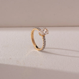 Angled view of oval diamond solitaire ring in yellow gold with a pavé diamond band, set on a soft neutral background, highlighting elevated setting and radiant sparkle.
